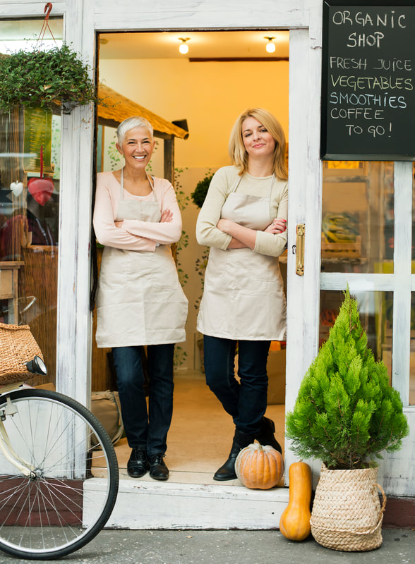 A pair of smiling women business owners, posing in aprons in front of their shop